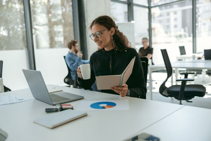 Women holds book and coffee while smiling at computer screen and sitting, colleagues in background 