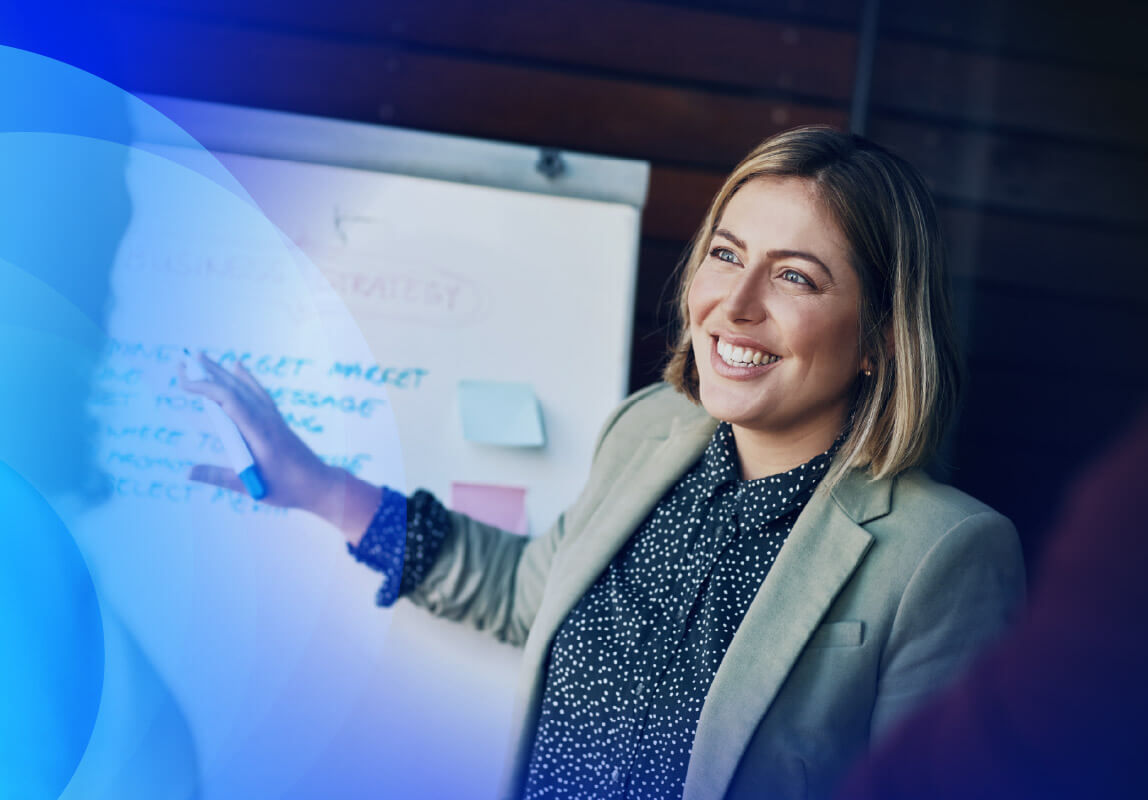 A smiling female professional in a light-colored blazer and polka dot shirt is standing in front of a whiteboard, pointing at it with a marker and presenting business strategy. 