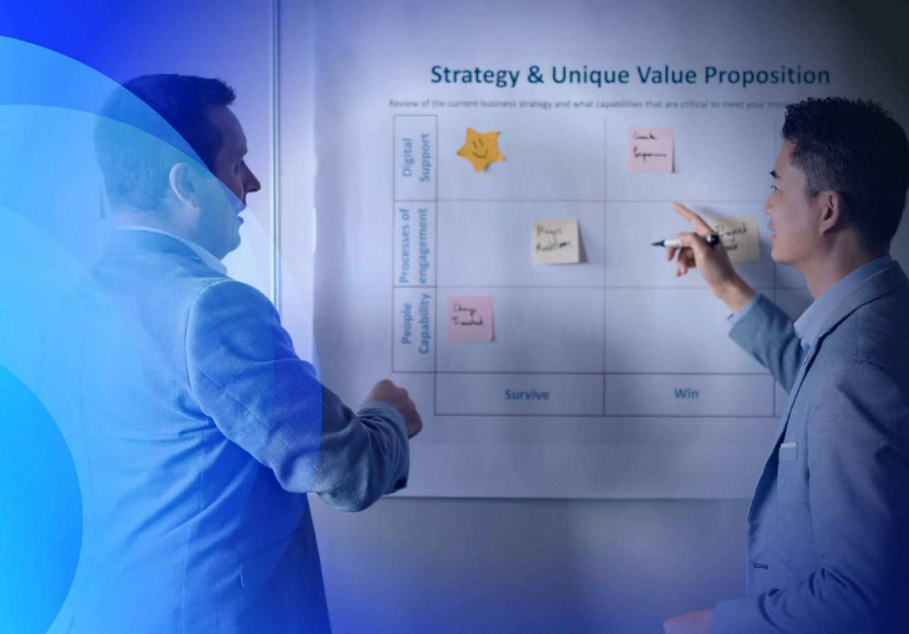 Two men writing on a sticky note stuck to a board