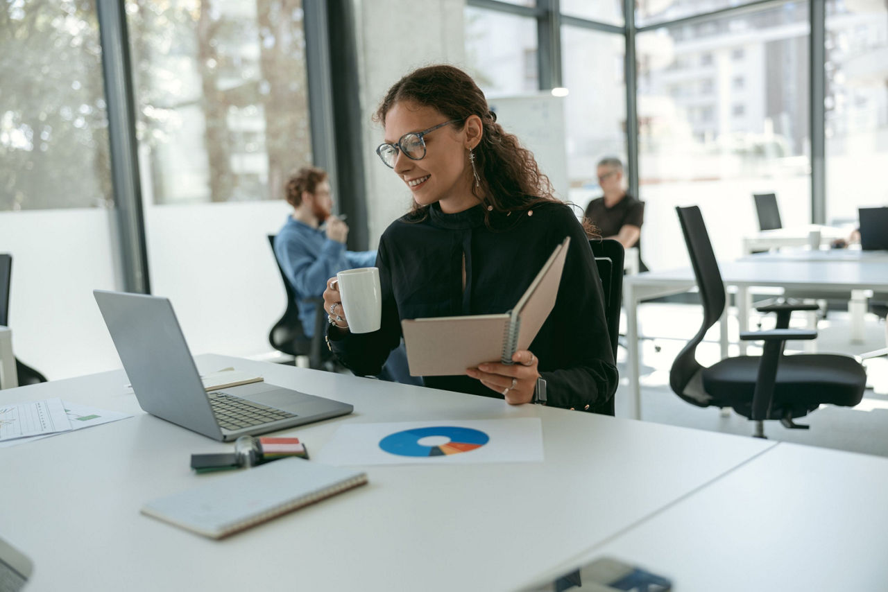 Women holds book and coffee while smiling at computer screen and sitting, colleagues in background 